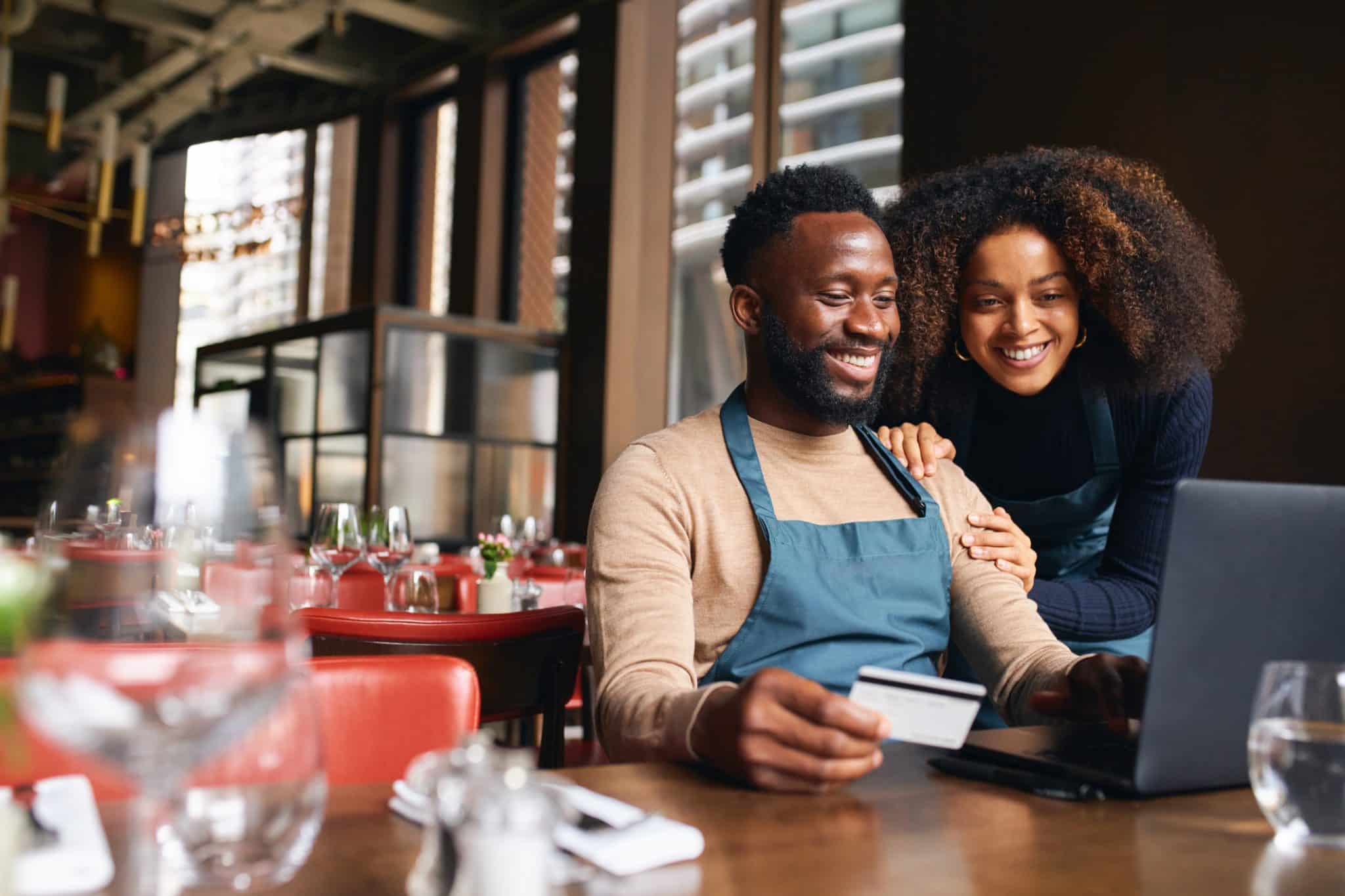 Two business owners sit in their restaurant, reviewing documents on their laptop as they prepare to apply for a business loan.